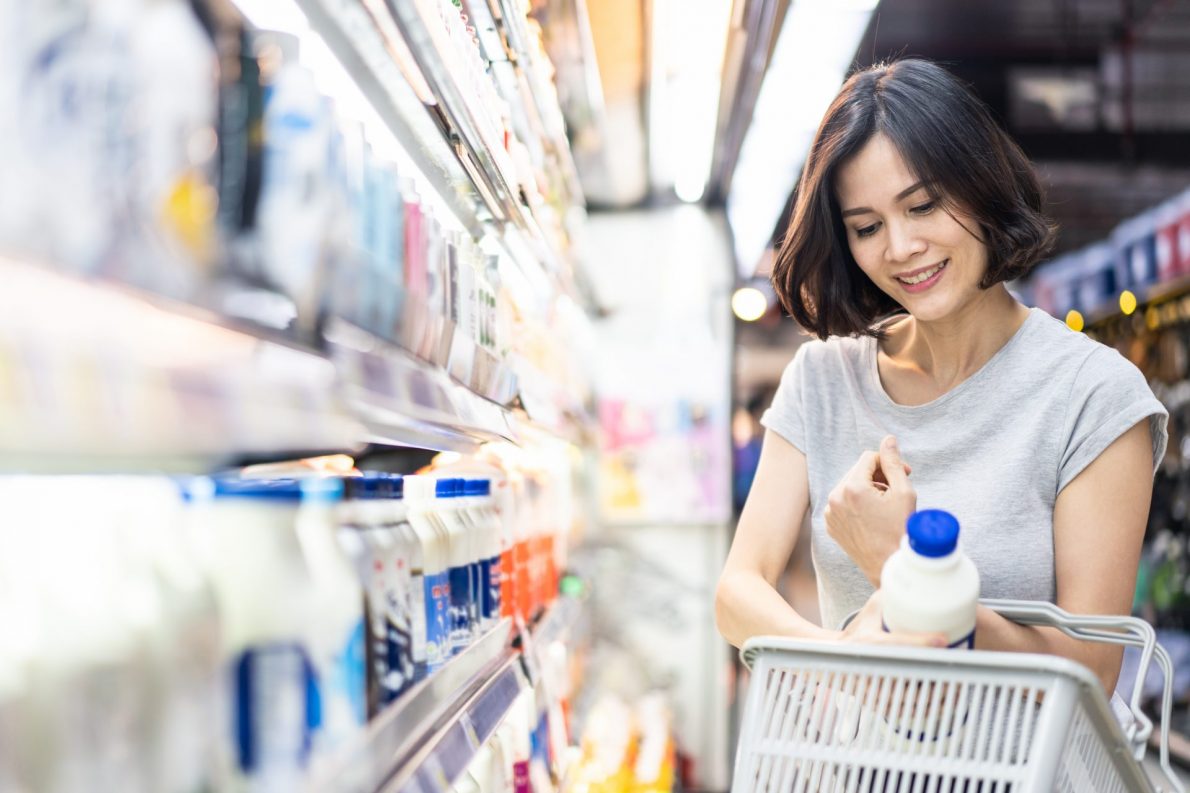 Young female consumer holding grocery basket, picking milk from shelf in supermarket